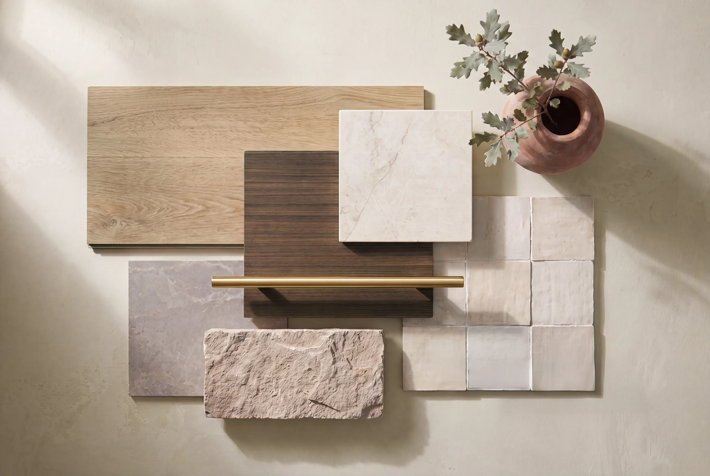 The CRE8R materials board: white oak flooring, walnut cabinetry, marble and quartz slabs, a brass bar pull, glazed cream tile, and rough limestone arranged and lit as a still life