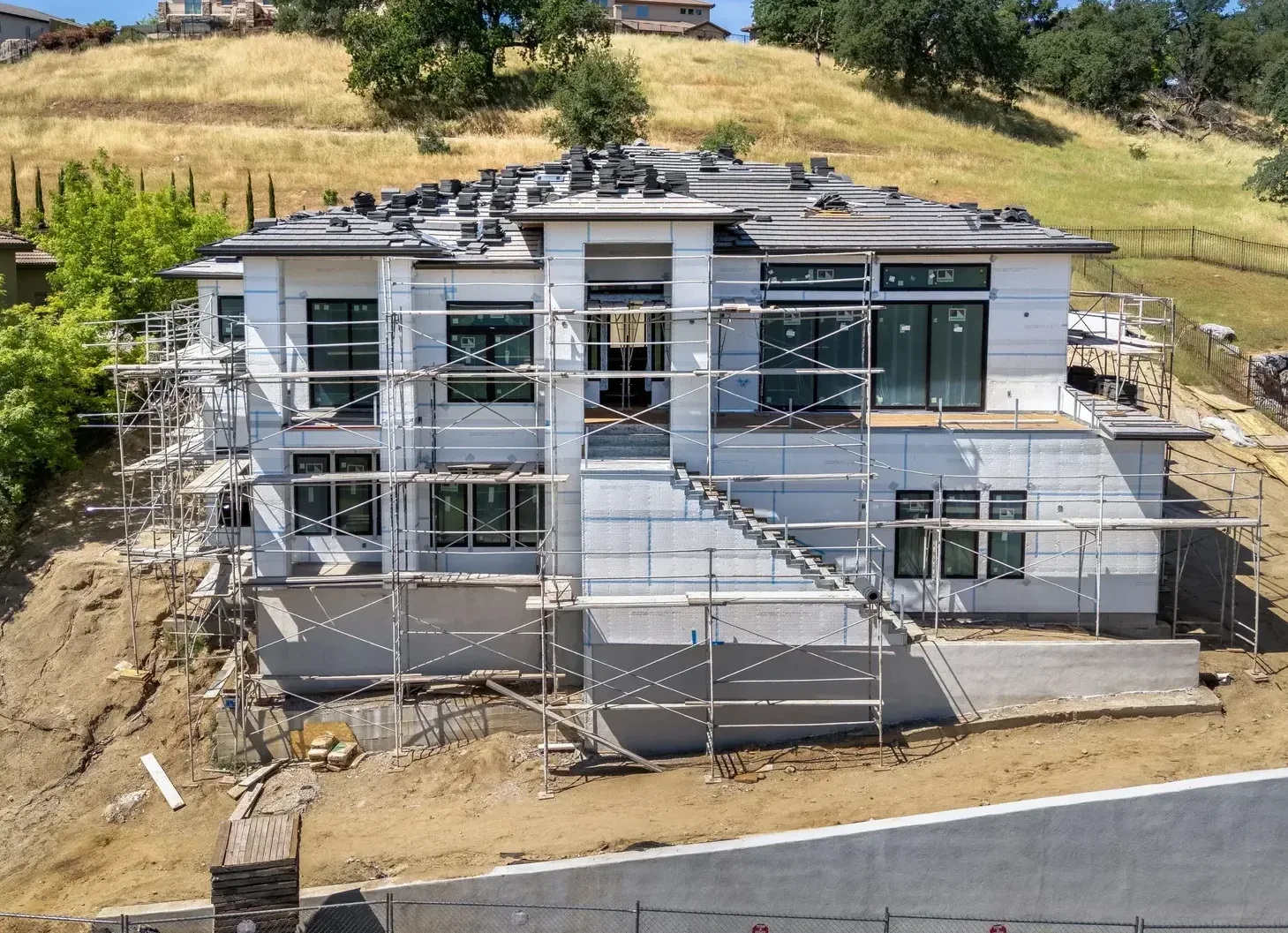 The front of 2578 Clubhouse Drive West in April 2026, head on: wrapped in scaffolding, roof tiles stacked along the ridge, bare stucco wrap and an unfinished stair