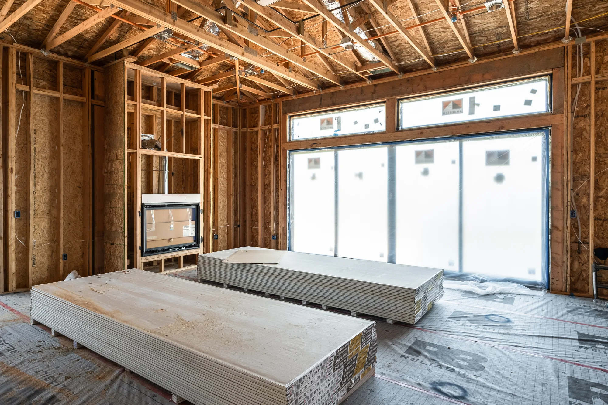The living room at framing stage: open studs, exposed trusses, the fireplace box set into its niche, and stacked drywall on the subfloor