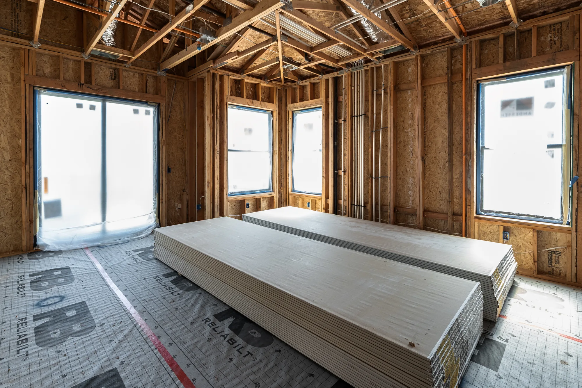 The primary bedroom at framing stage: open studs, a tray ceiling roughed into the trusses, a slider opening and two window openings sheeted over