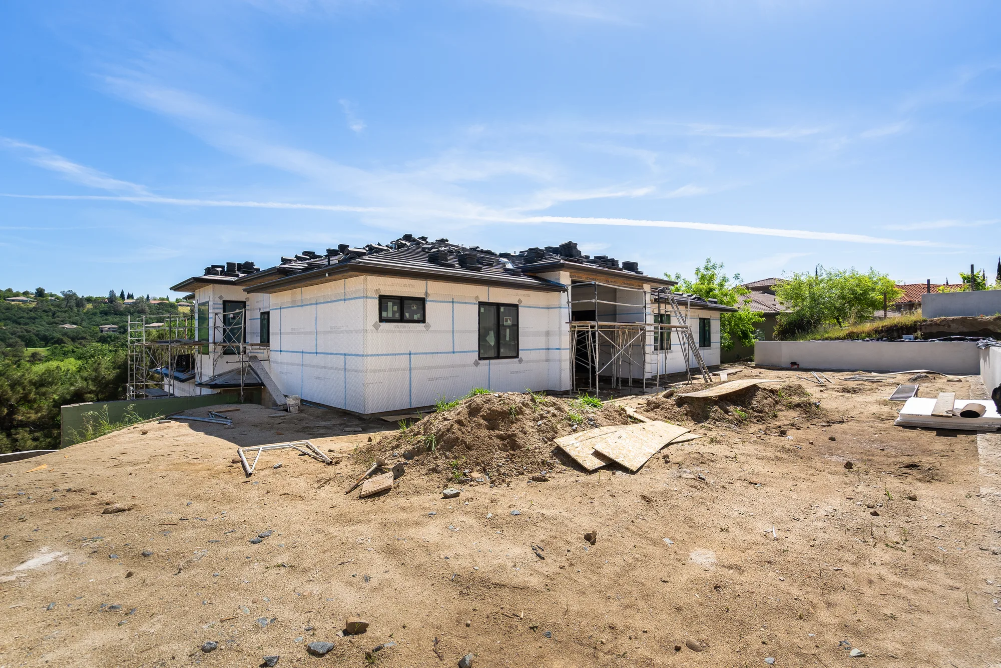 The rear of 2578 Clubhouse Drive West in April 2026: bare stucco wrap, roof tiles stacked along the ridge, and a graded dirt lot where the pool would go