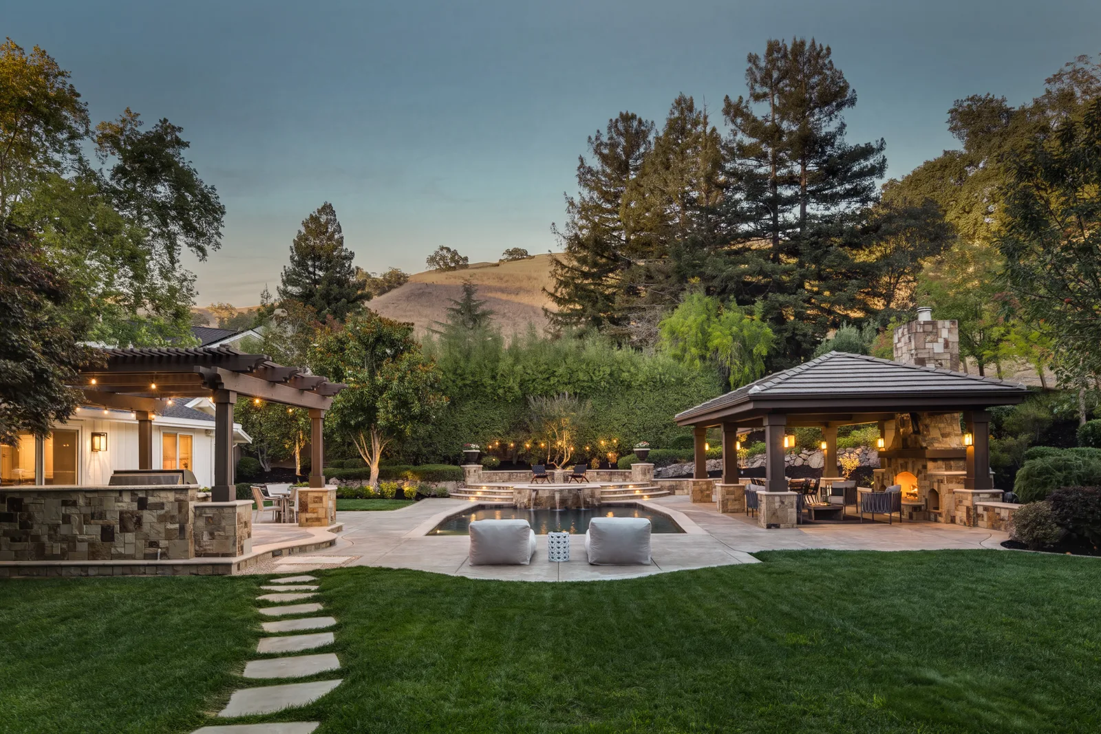 The grounds at 2780 Mossy Oak Drive at dusk: pool and spa, a stone pavilion with a lit fireplace, pergola, and lawn against the hills