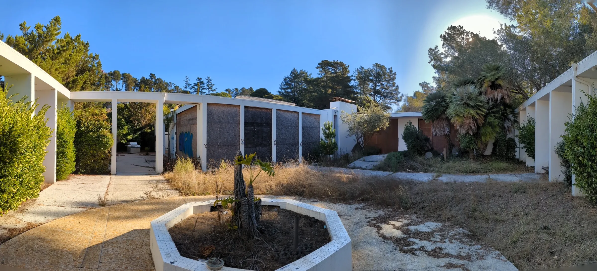 The courtyard at 5645 Bacon Road in 2023: boarded, X-marked window bays, dry weeds, and a dead planter