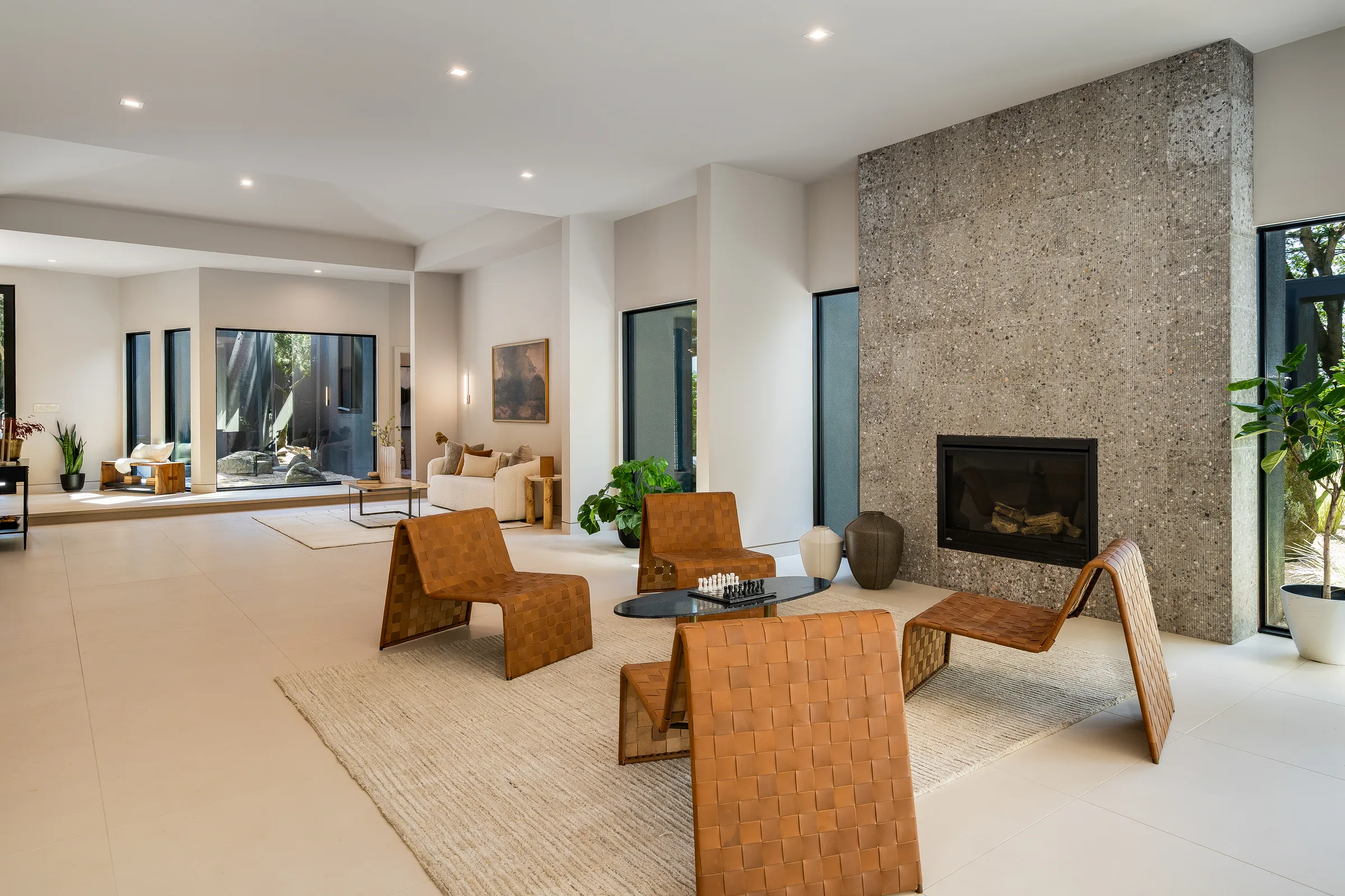 A modern living room: a full-height aggregate stone fireplace wall, woven leather lounge chairs on a wool rug, and floor-to-ceiling black-framed windows opening to trees.