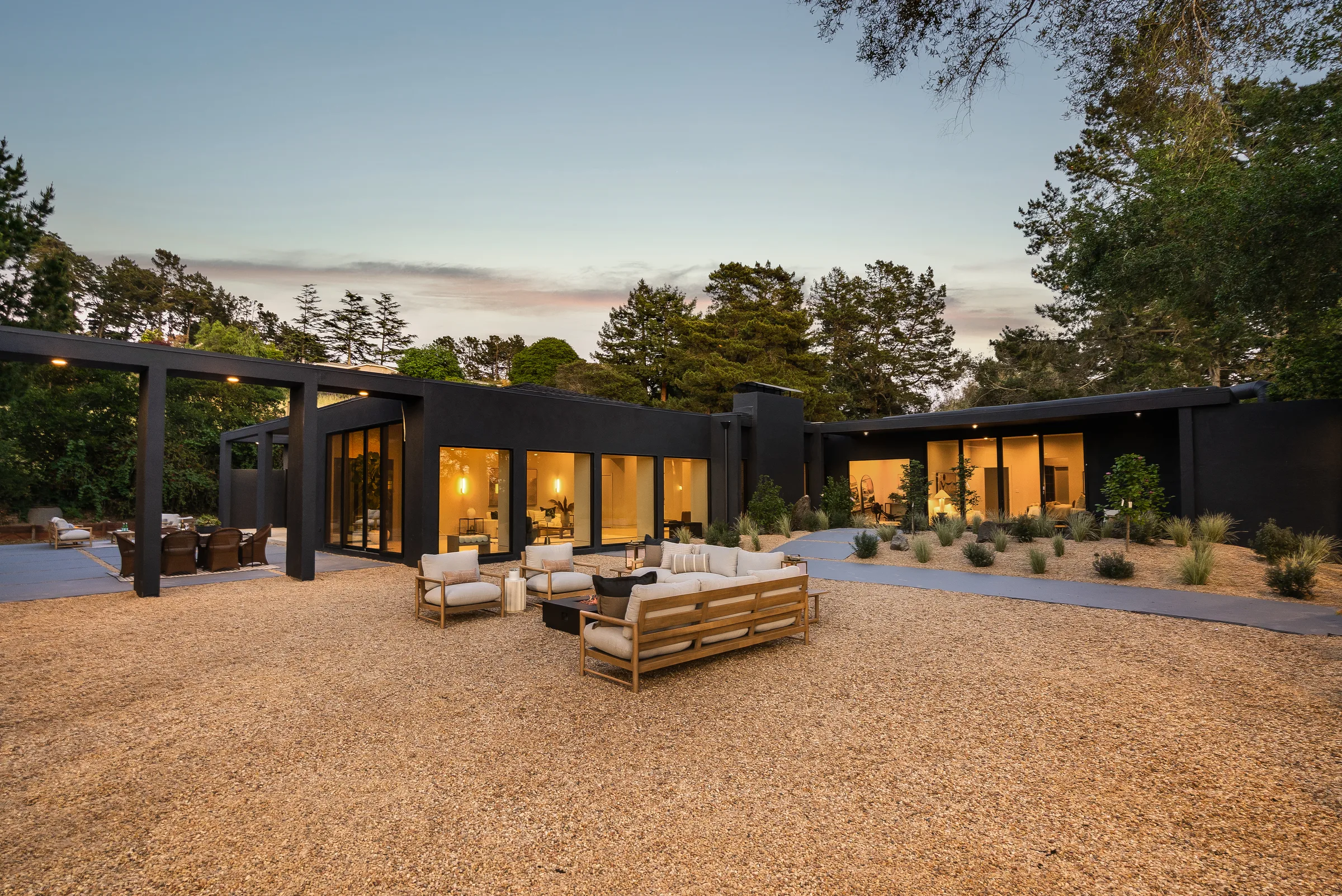The same courtyard at dusk after the rebuild: black stucco, glowing glass bays, gravel court, and a fire-pit seating area