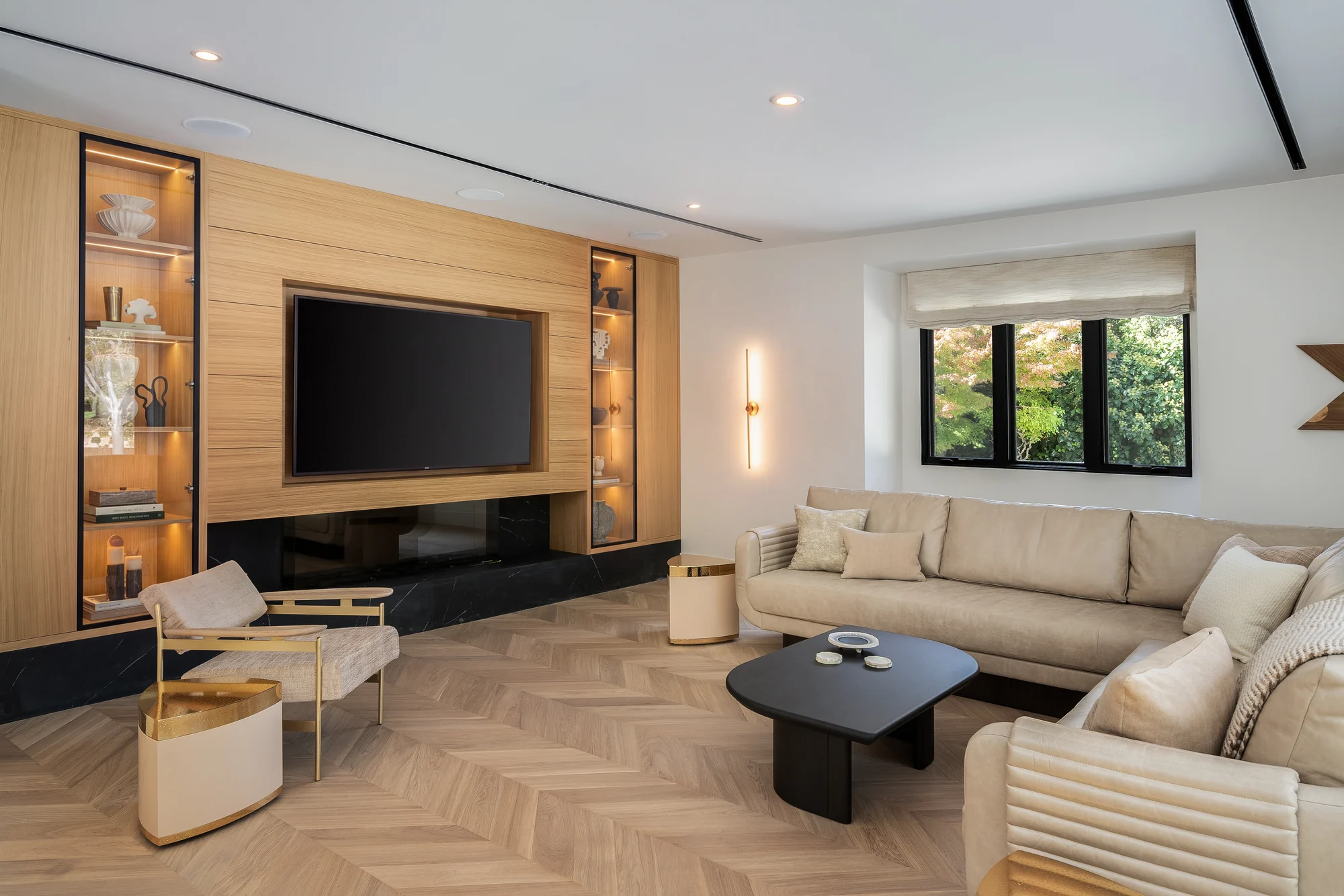 A finished living space in a Silicon Valley residence: a wall-mounted screen set into white oak millwork between lit display cabinets, a recessed hearth in black marble below, herringbone floors and low cream seating
