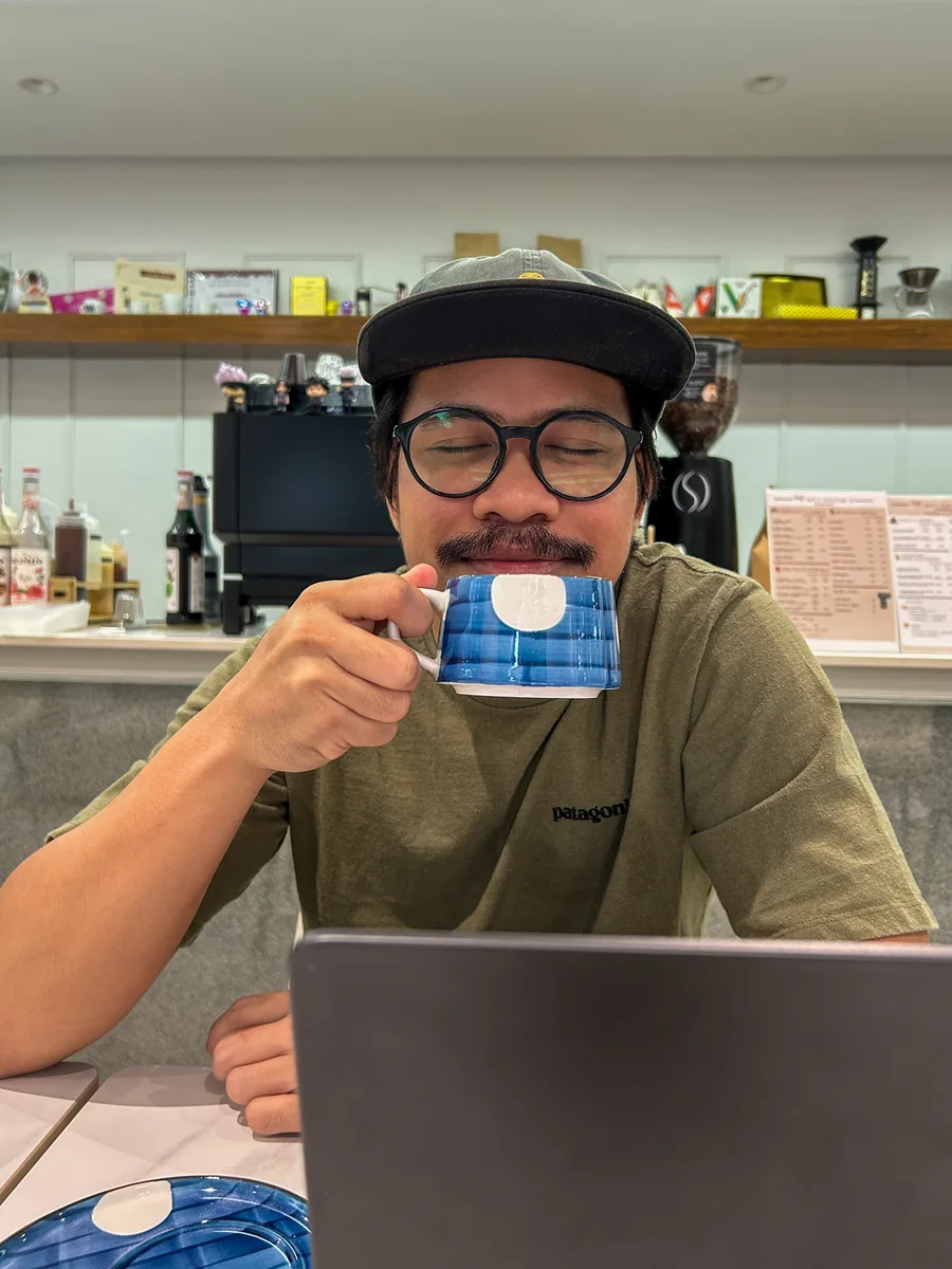 Nico at a café counter with a laptop, eyes closed, sipping from a mug.