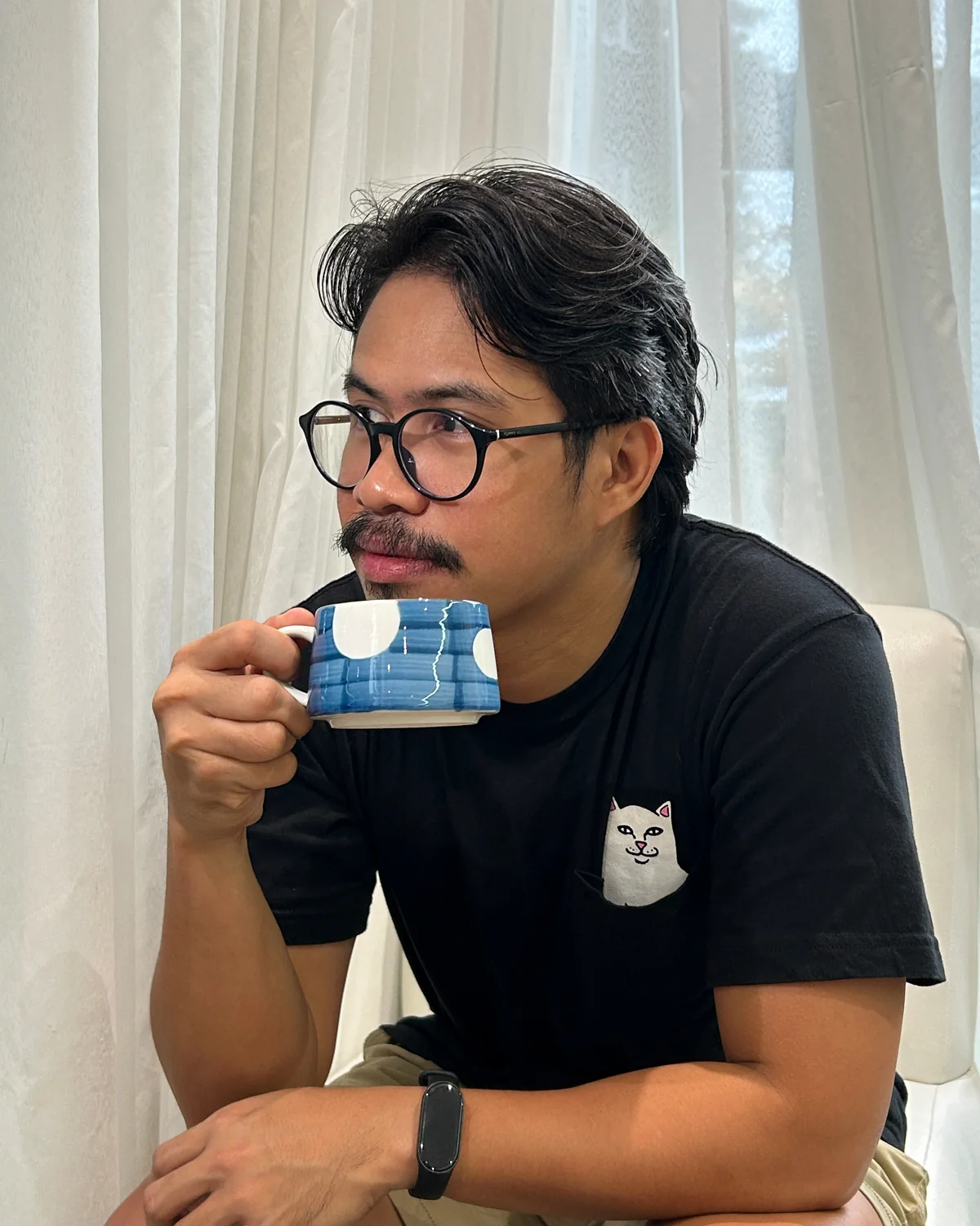 Nico Palma in a black tee and glasses, drinking from a blue mug by the window.